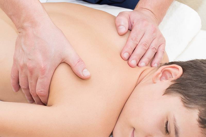 Ein lächelnder Junge liegt entspannt auf einer Massageliege im Arkona Strandhotel Rügen, während eine Rückenmassage durchgeführt wird.