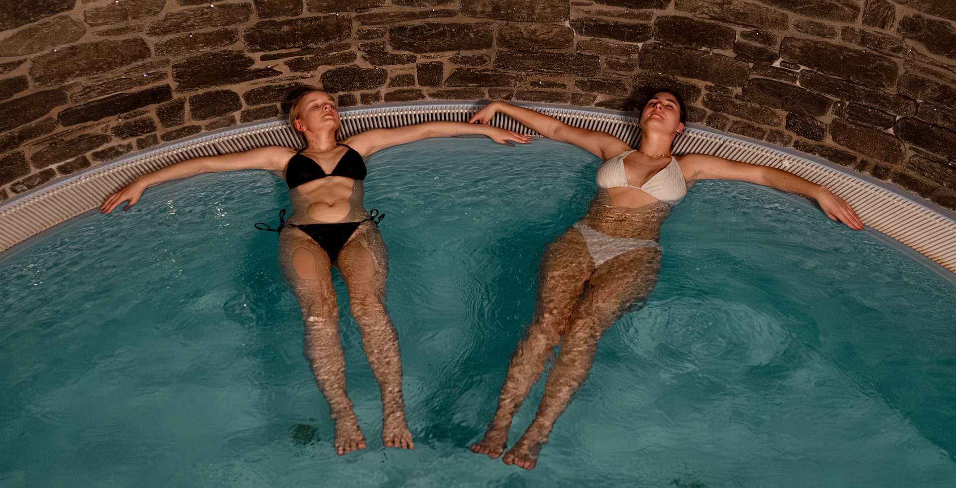 Two women relax next to each other in the warm wellness pool with natural stone wall at the Rugard Thermal Standhotel on Rügen.